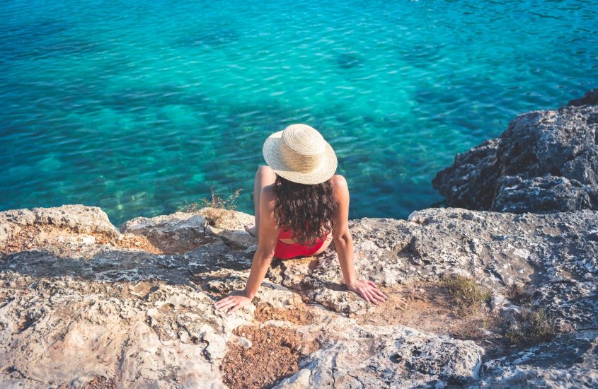 woman in hat and red bikini sitting on rock near water