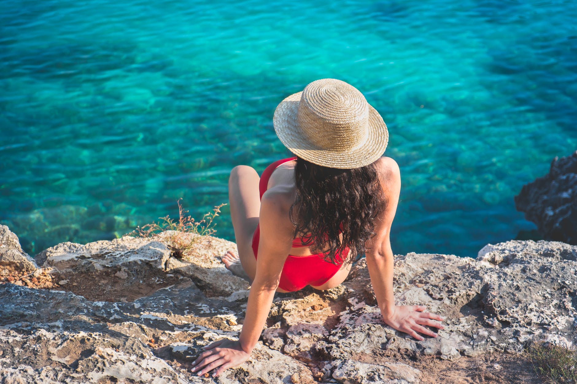 woman in red bikini wearing straw hat sitting on rock near water